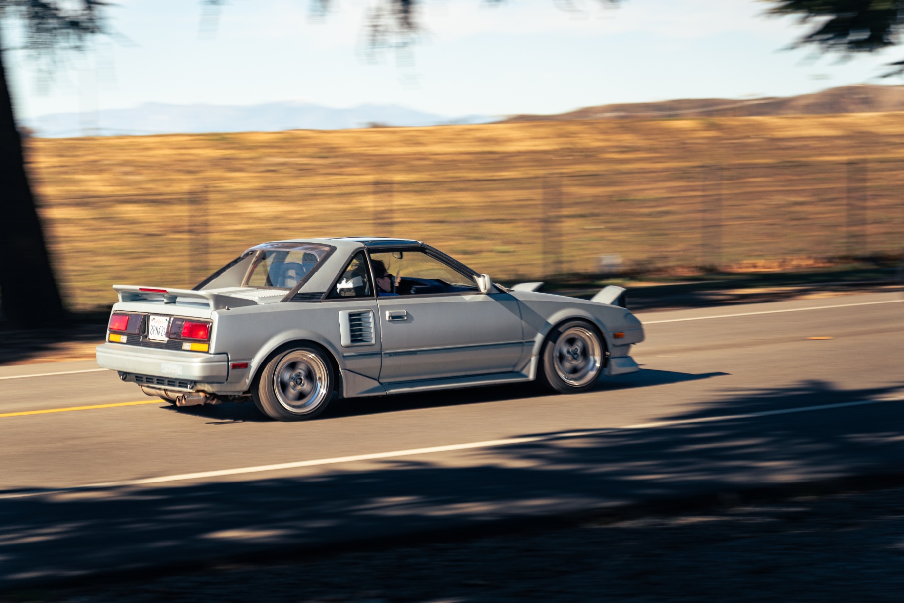 Silver Toyota MR2 AW11 with pop-up headlights panning past the hills