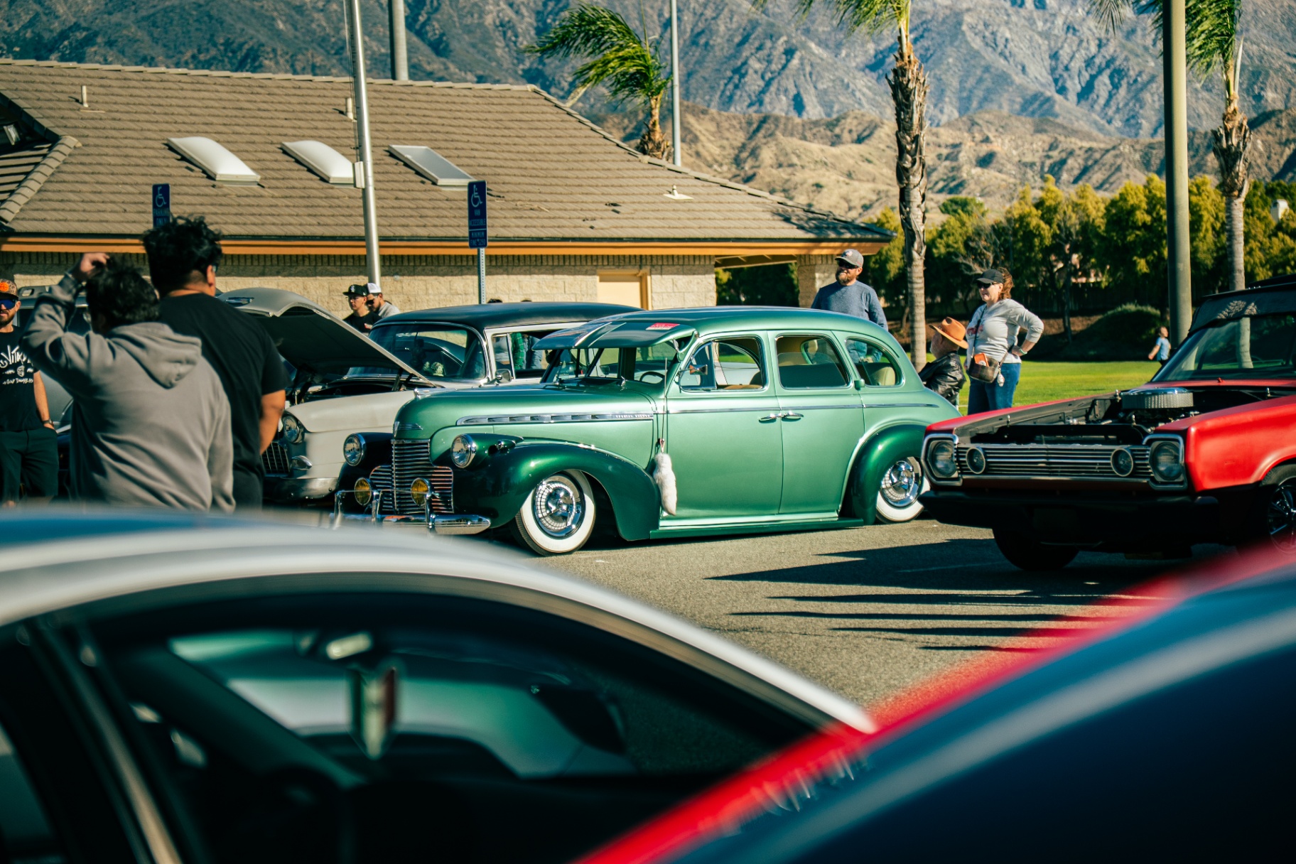 Bagged green vintage Chevy sedan among lowriders with mountain backdrop