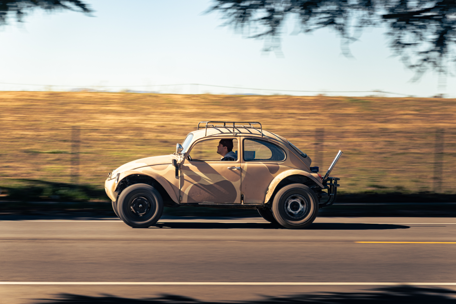 Tan VW Beetle with roof rack and desert camo paint cruising past golden hills